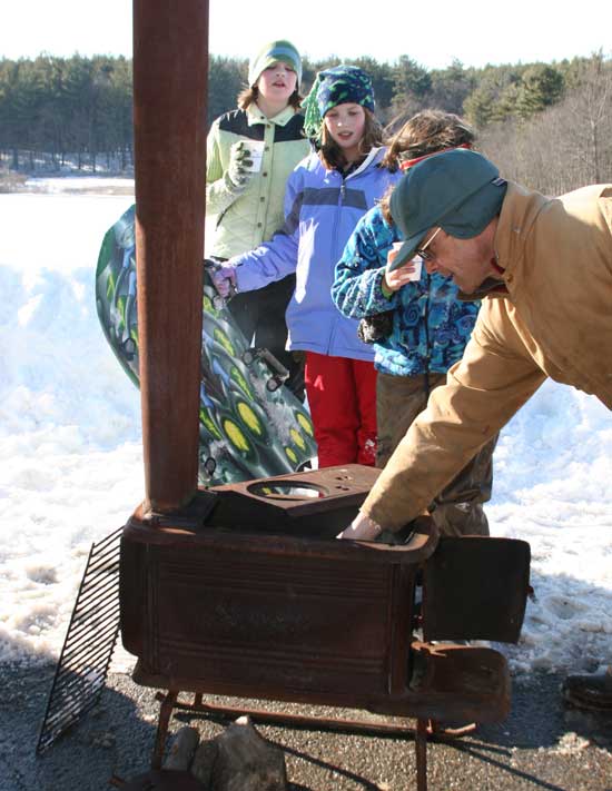 Sarah Eldredge and Madeline Hettich watch as Bob Donaldson stokes his portable woodstove to keep the hot drinks coming at his daughter's sledding birthday party.