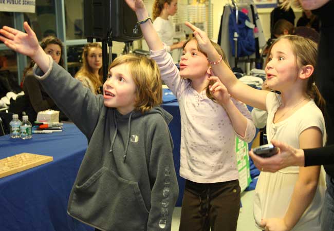 Gabe Sennott, Leila Grant, and Madison Drummey all reach for the microphone so that they can take a turn announcing the numbers in bingo during a family game night at Bromfield on Friday, March 13.