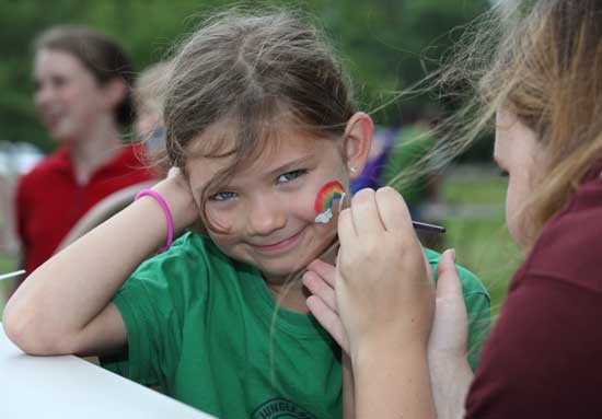 Isabelle Planchet smiles while Anna Fellows paints a rainbow on her cheek.