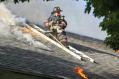 Firefighter Charles Dillon of Ayer pauses from chainsawing to survey the scene as flames lap out of the roof below. He cut through the roof of the Harvard Historical Society in Still River after a blaze broke out Tuesday morning.
