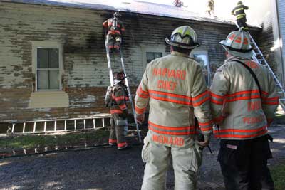 Firefighter Charles Dillon of Ayer pauses from chainsawing to survey the scene as flames lap out of the roof below. He cut through the roof of the Harvard Historical Society in Still River after a blaze broke out Tuesday morning.