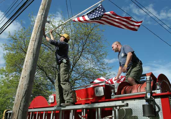 Bill Barton and Bill McElhaney place flags on telephone poles from atop engine #4 in preparation for the Memorial Day parade.