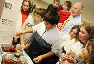 Xavier Neang, center, puts a Mentos candy into a bottle of Coke to show the effect of carbon dioxide buildup in a small space. Left to right are Jane Sullebarger, Pat Pesa, Phoebe Carona, sixth-grader Xavier Neang, Charlotte O’Shea, Jim O’Shea, Elly vandeVaart, Jillian Carr, and Eliza Agosta.